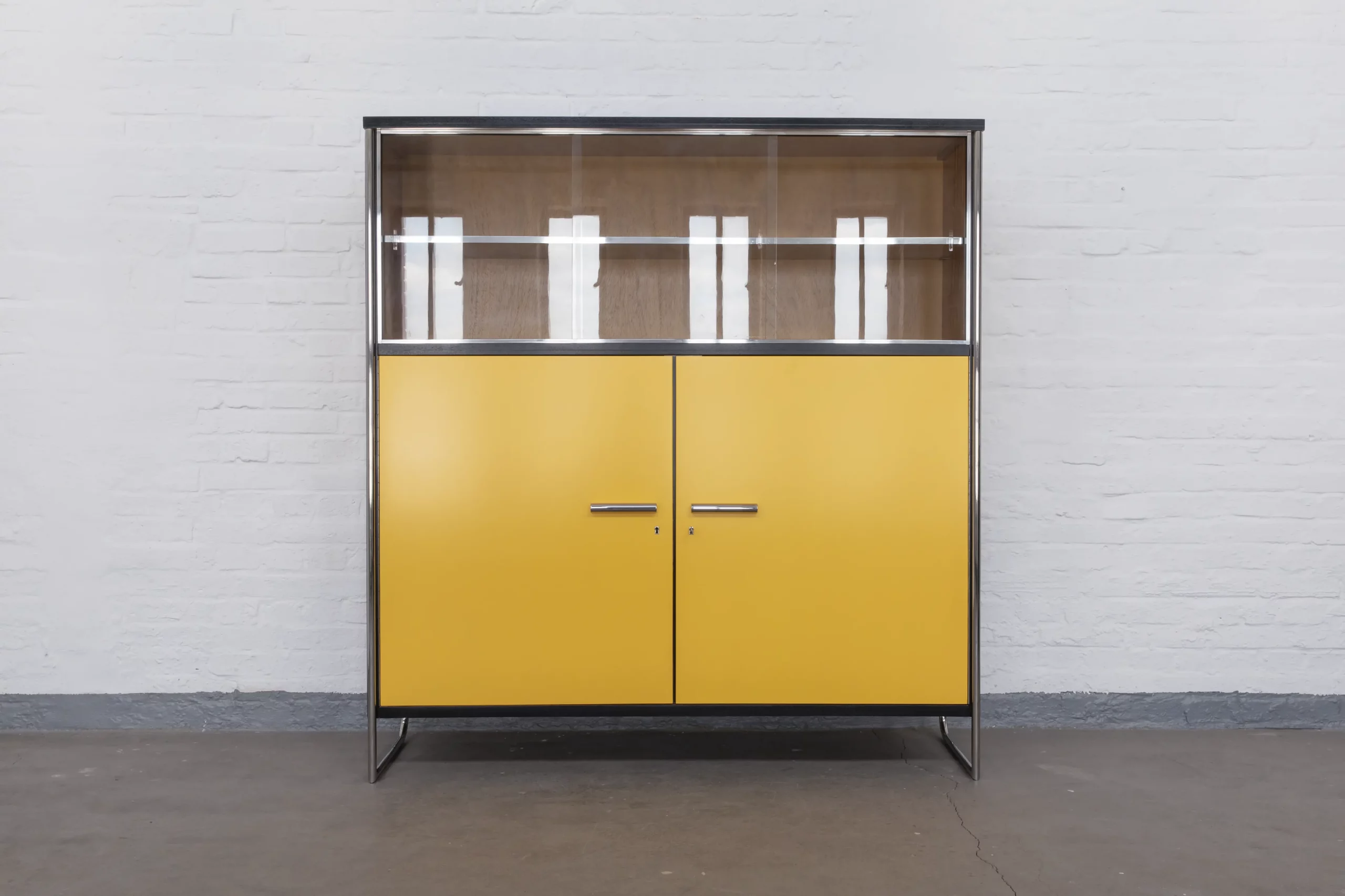 Display cabinet in the style of Bauhaus Modernism with polished tubular steel frame, sliding glass doors, yellow veneered wood panels and chrome-plated details.